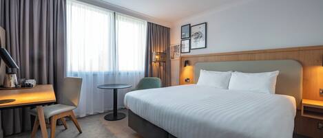 Habitació d'hotel amb un llit gran, una taula de treball amb una cadira, una taula petita i una làmpada.
