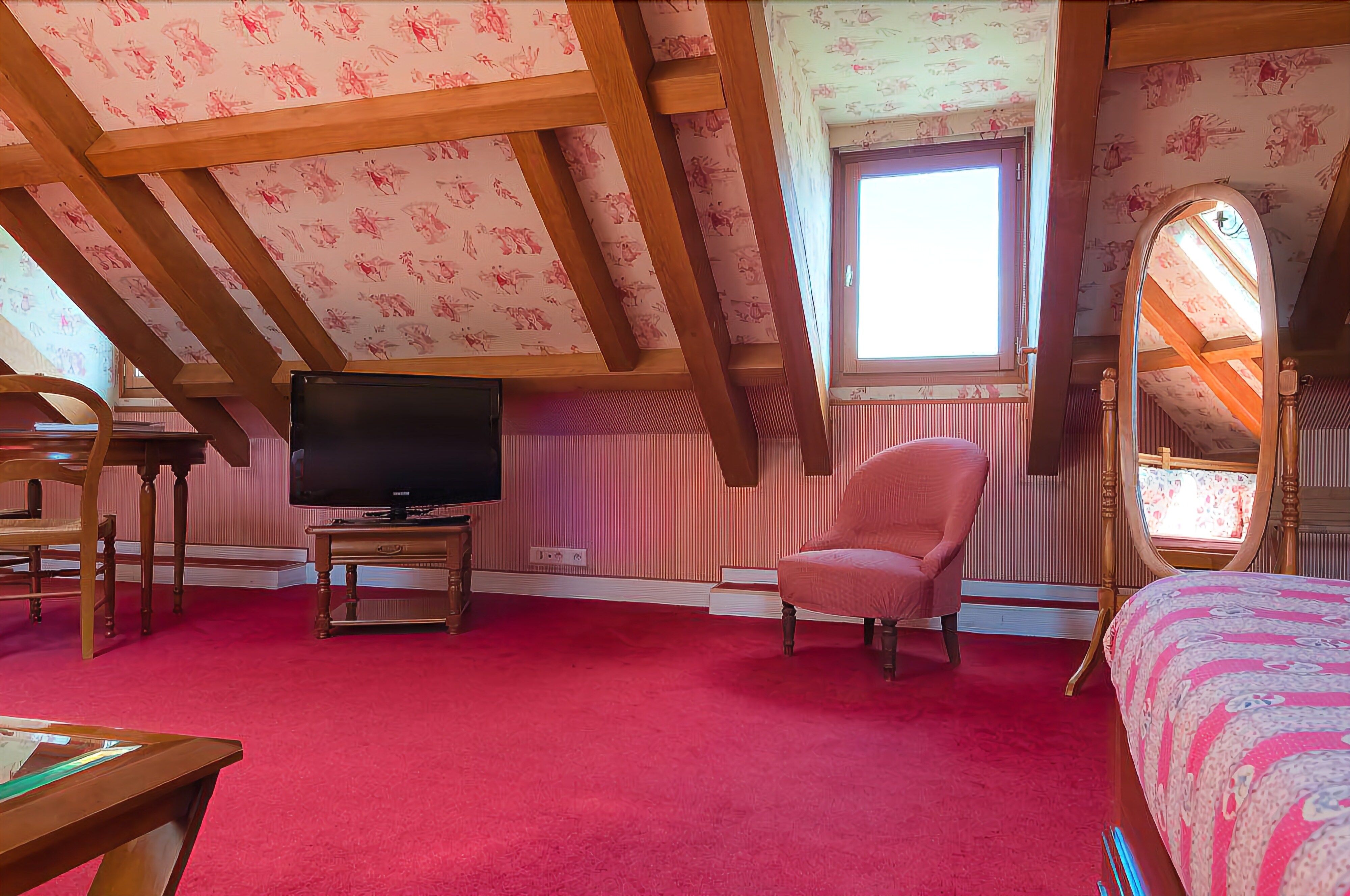 A bedroom with a sloped ceiling, a television, a pink armchair, a bed with a patterned bedspread, and a large mirror.