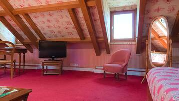 A bedroom with a sloped ceiling, a television, a pink armchair, a bed with a patterned bedspread, and a large mirror.