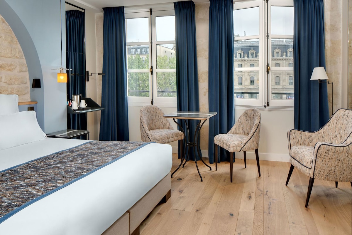 A hotel room with a bed, a table, two chairs, a lamp, and a view of buildings through large windows.