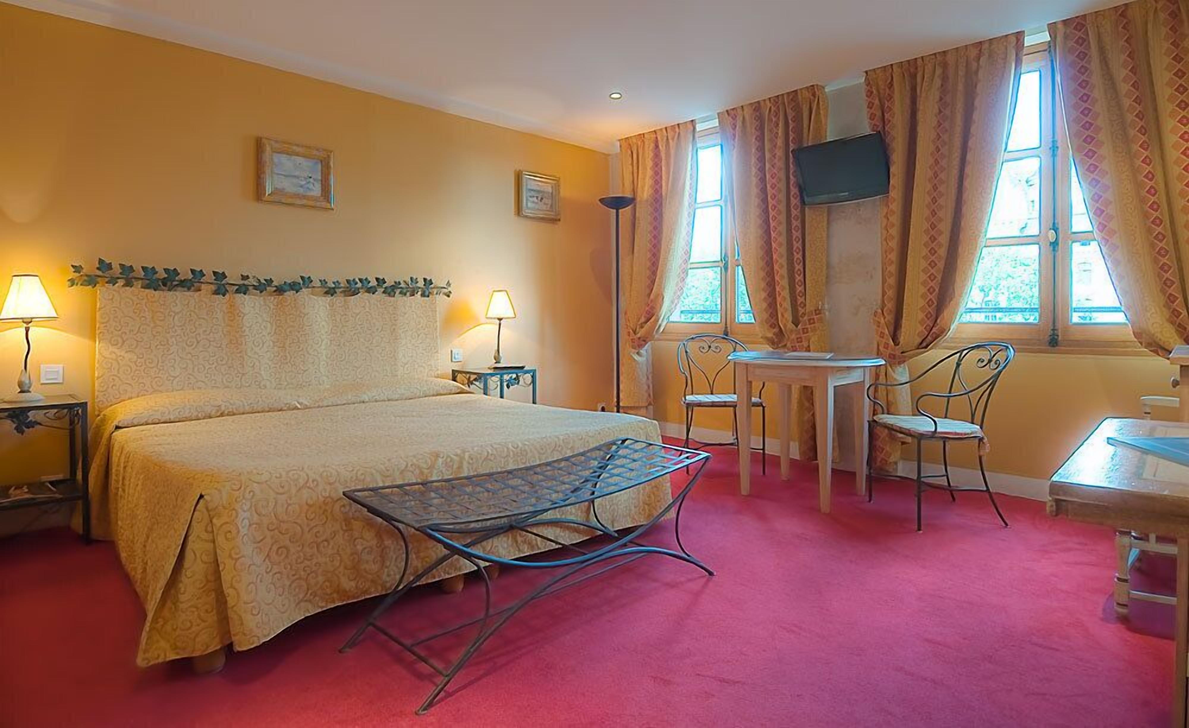 Habitación de hotel con cama, dos mesitas de noche, escritorio con silla, pequeña mesa con otra silla y televisor fijado en la pared.