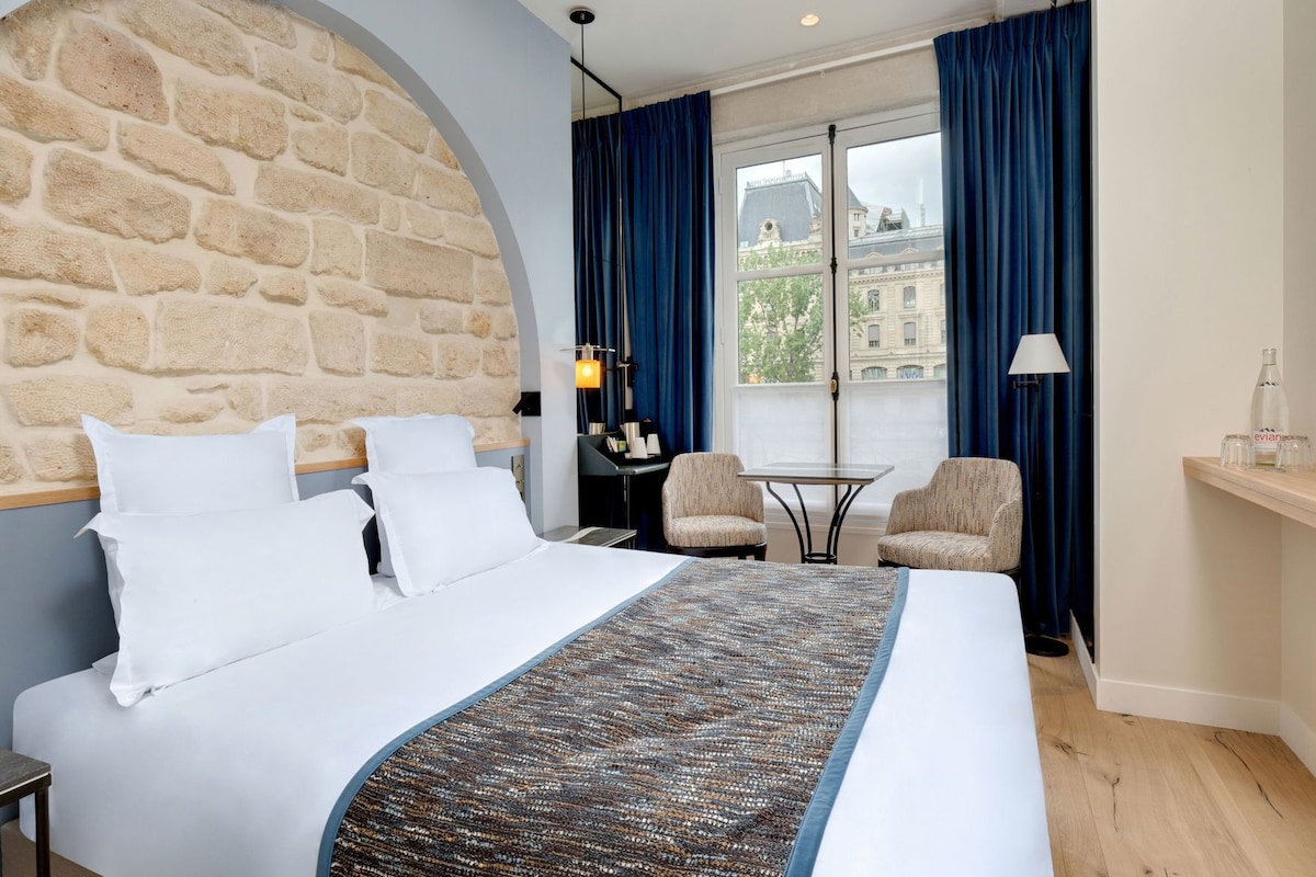A hotel room with a stone wall, a bed with white linens, a small table, two armchairs, and a view of a building through a window.