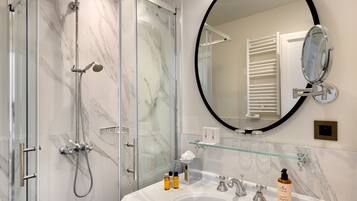 Un baño moderno con ducha de vidrio, espejo redondo y lavamanos blanco con artículos de aseo.