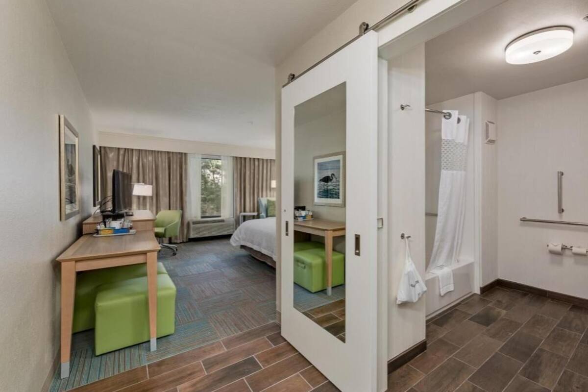 A hotel room with a bed, a desk, a green ottoman, a shower, and a bathroom with a towel rack.