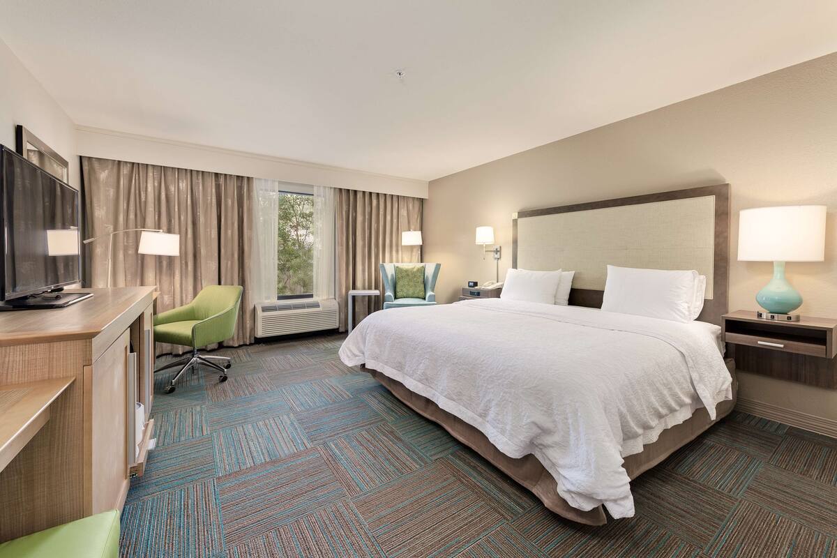 A hotel room with a large bed, a desk with a television, a green chair, and a window with curtains.