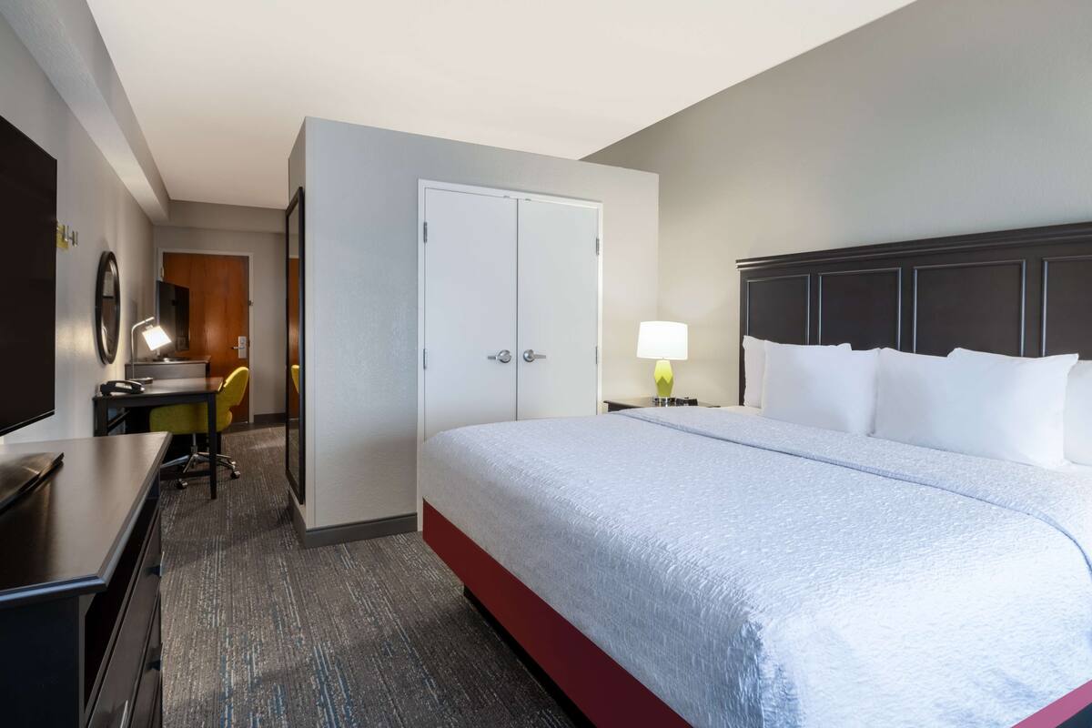 Een hotelkamer met een bed, een bureau, een stoel, een bureaulamp, een nachtkastje met lamp en een kledingkast.