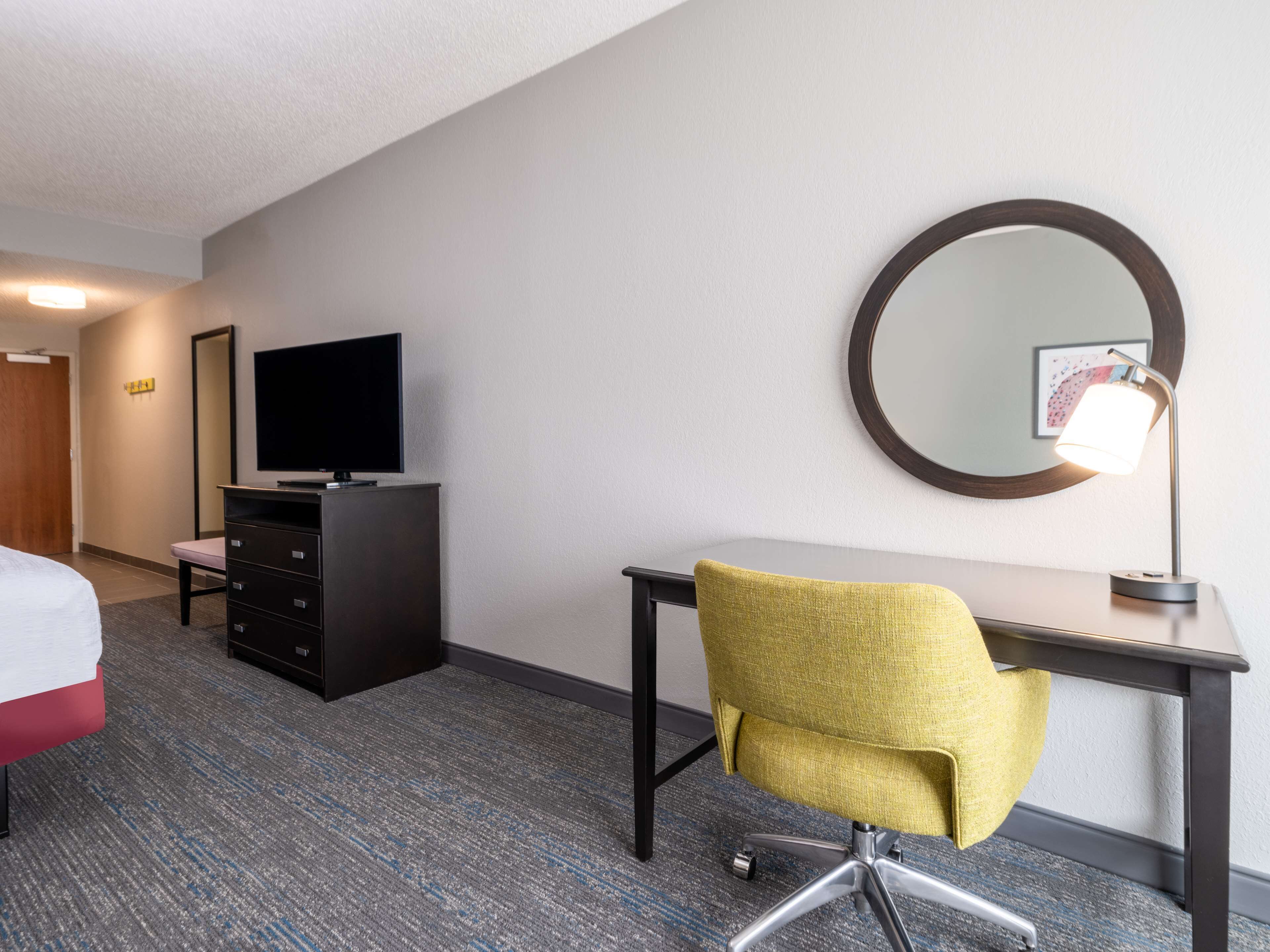 Habitación de hotel con cama, escritorio, silla, televisor, cómoda, espejo y lámpara.
