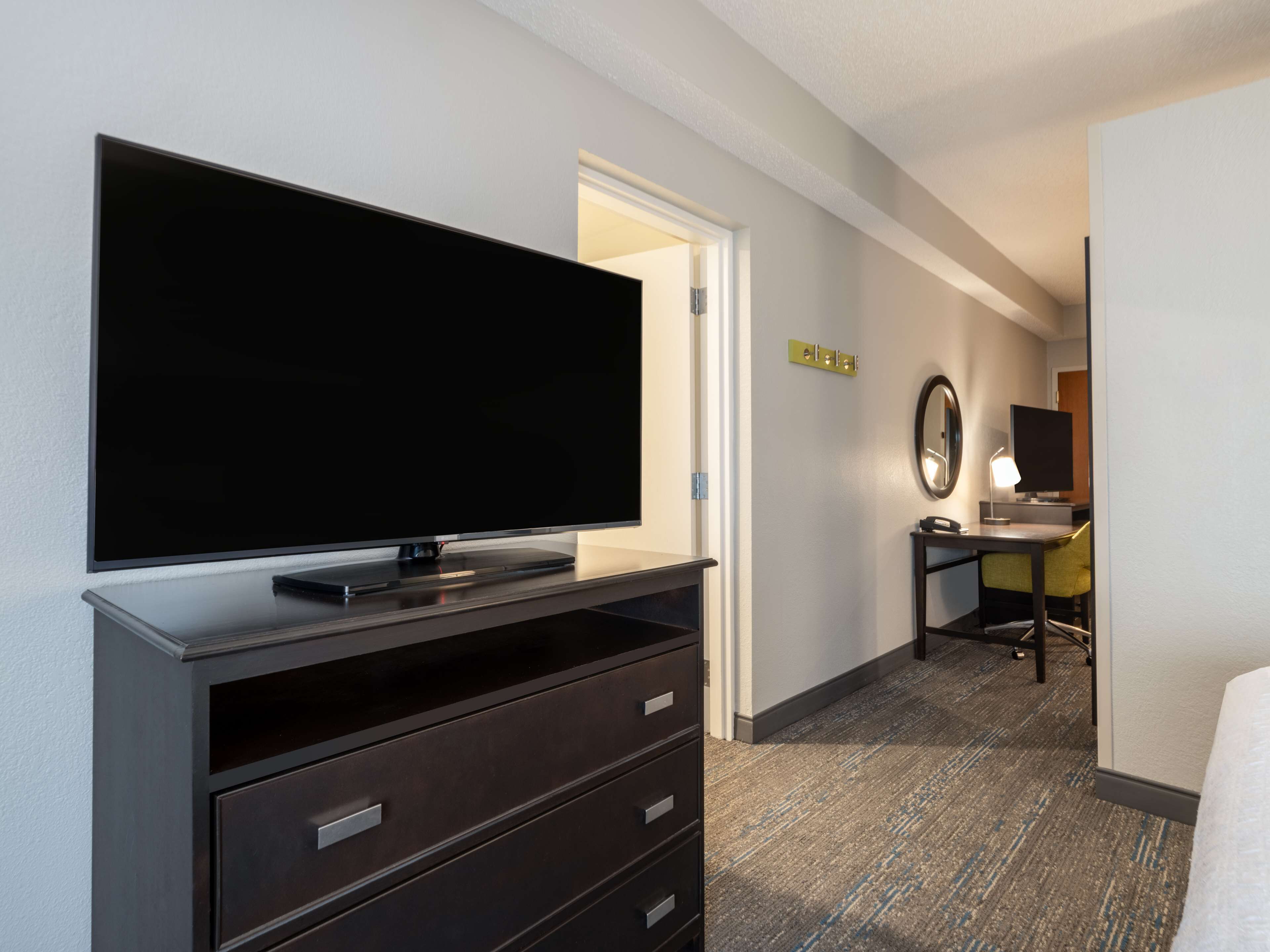 Ein Hotelzimmer mit Flachbildfernseher auf einer Kommode, einem Schreibtisch mit Stuhl und einem kleinen Tisch mit Lampe.