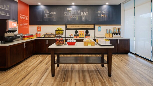 A modern kitchen area with a wooden table, a coffee machine, and various food items displayed on the counter. - Hampton Inn West Palm Beach Florida Turnpike (West Palm Beach)