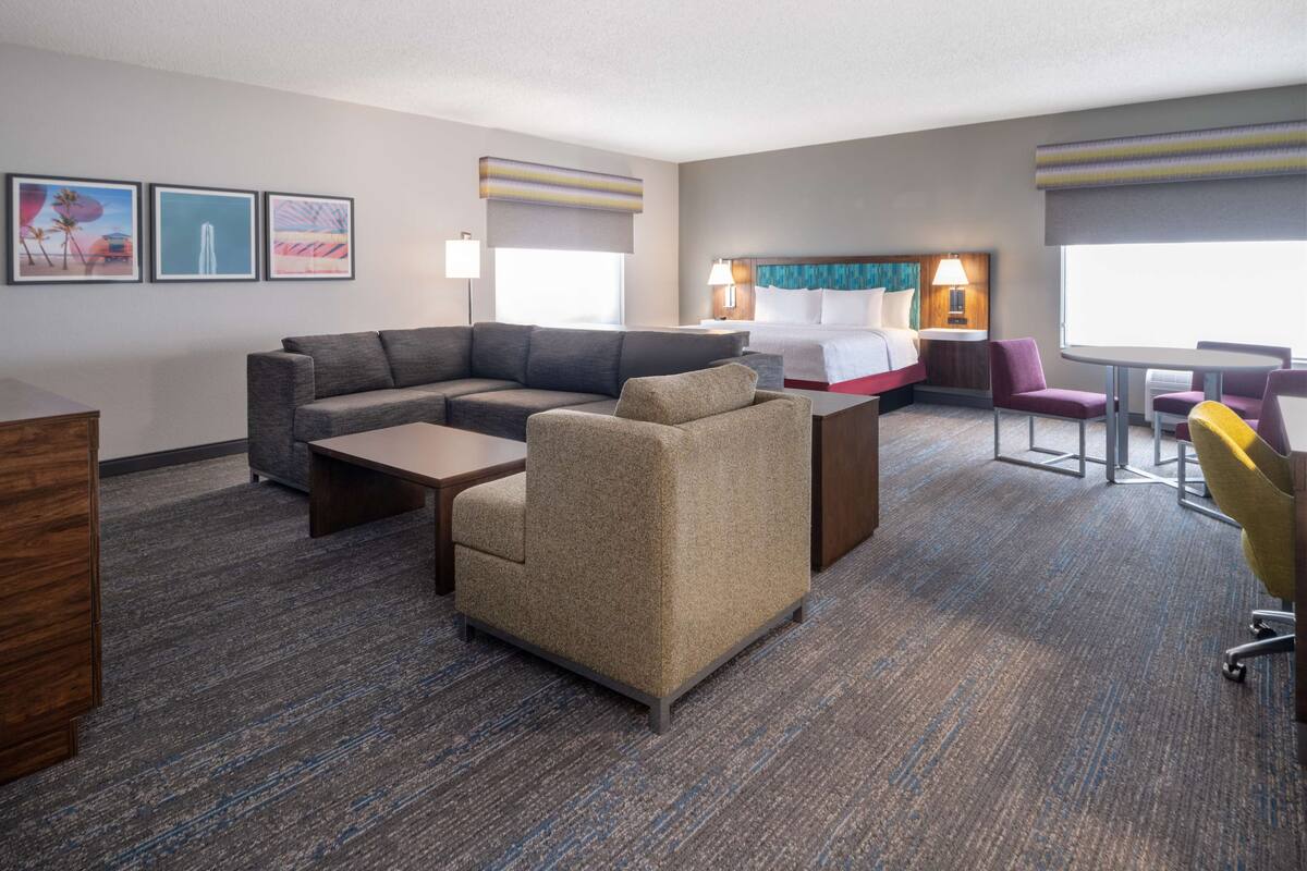 Een hotelkamer met een modulaire bank, een fauteuil, een tafeltje, een bed met hoofdeinde, een bureau en een stoel. Er hangen prestaties aan de muur, een raam met jaloezieën en een vloerkleed.