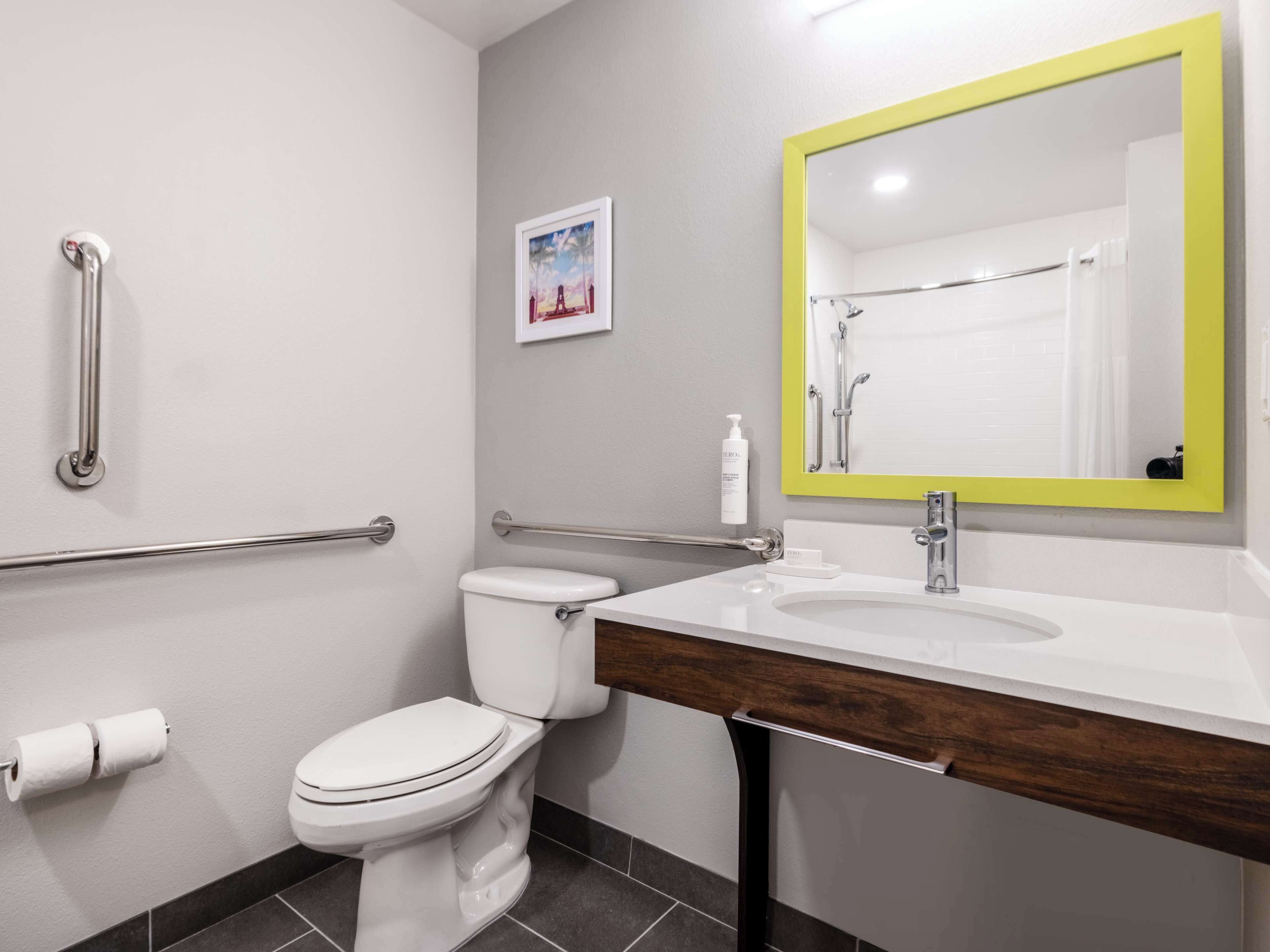 A modern bathroom with a white toilet, a sink with a wooden countertop, a mirror with a yellow frame, and a handrail.