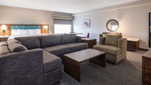 A modern hotel room with a large sectional sofa, a smaller armchair, a coffee table, and a desk with a chair. There is a window with a view, a wall-mounted lamp, and a framed picture. - Hampton Inn West Palm Beach Florida Turnpike (West Palm Beach)