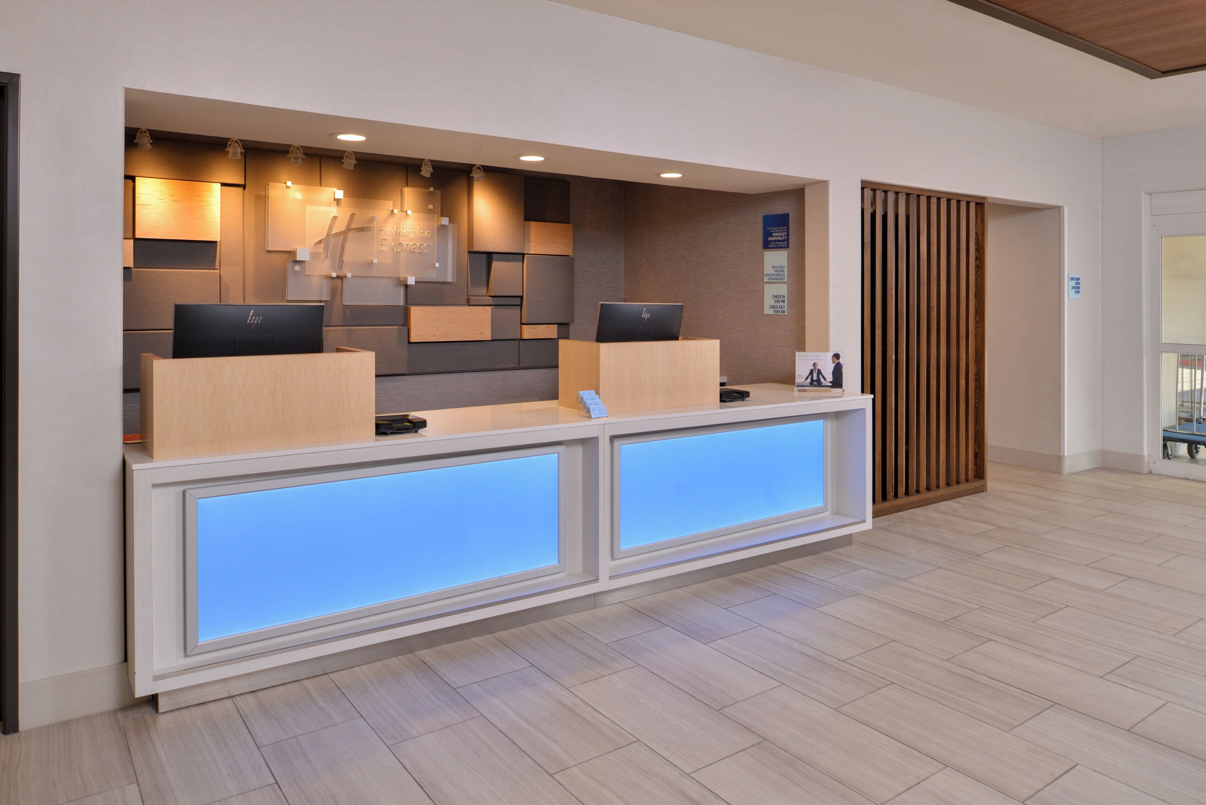 Un mostrador de recepción con dos monitores de computadora, un panel de pared de madera y un panel iluminado en azul.
