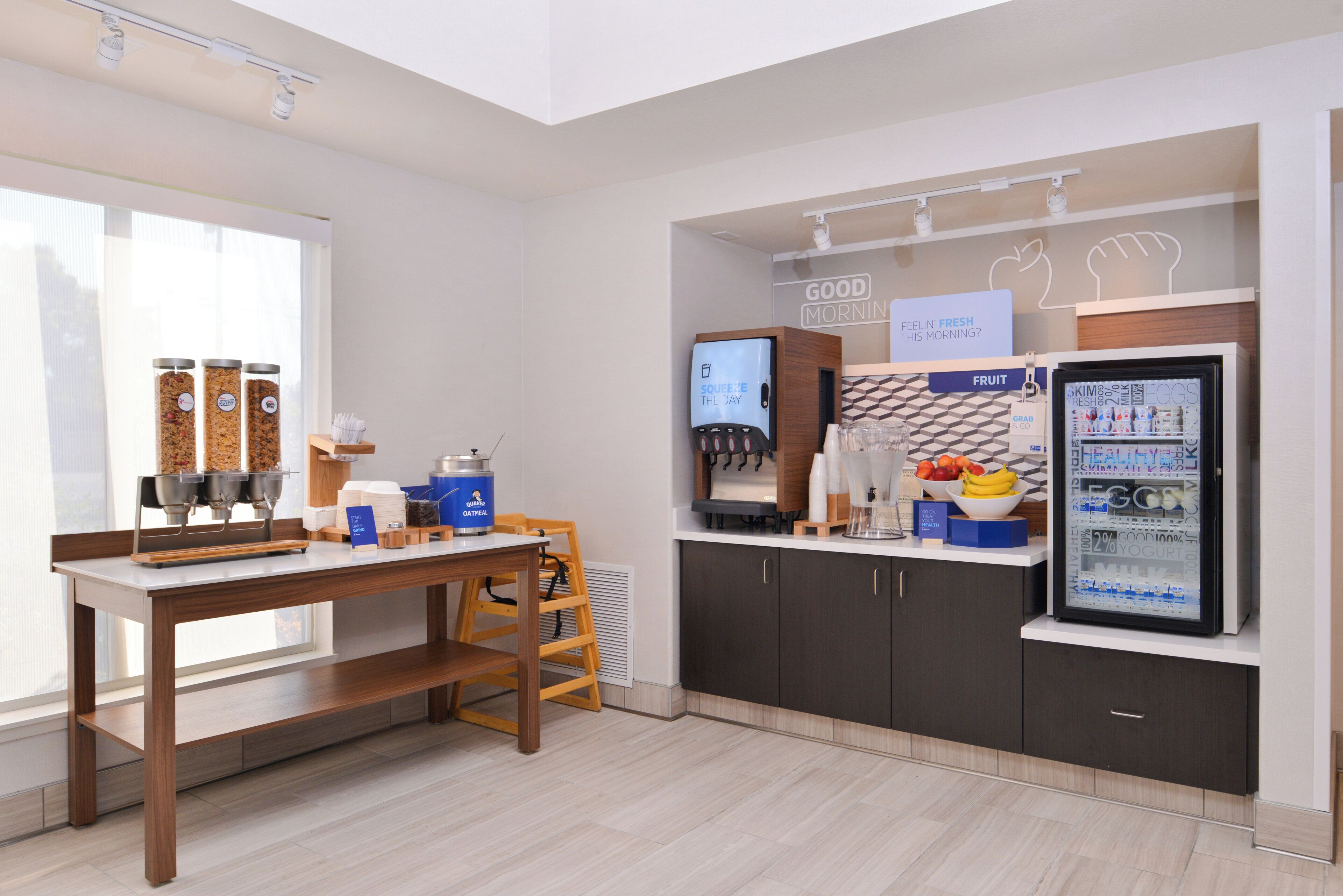 A modern kitchenette with a coffee machine, a fruit dispenser, and a refrigerator stocked with beverages.