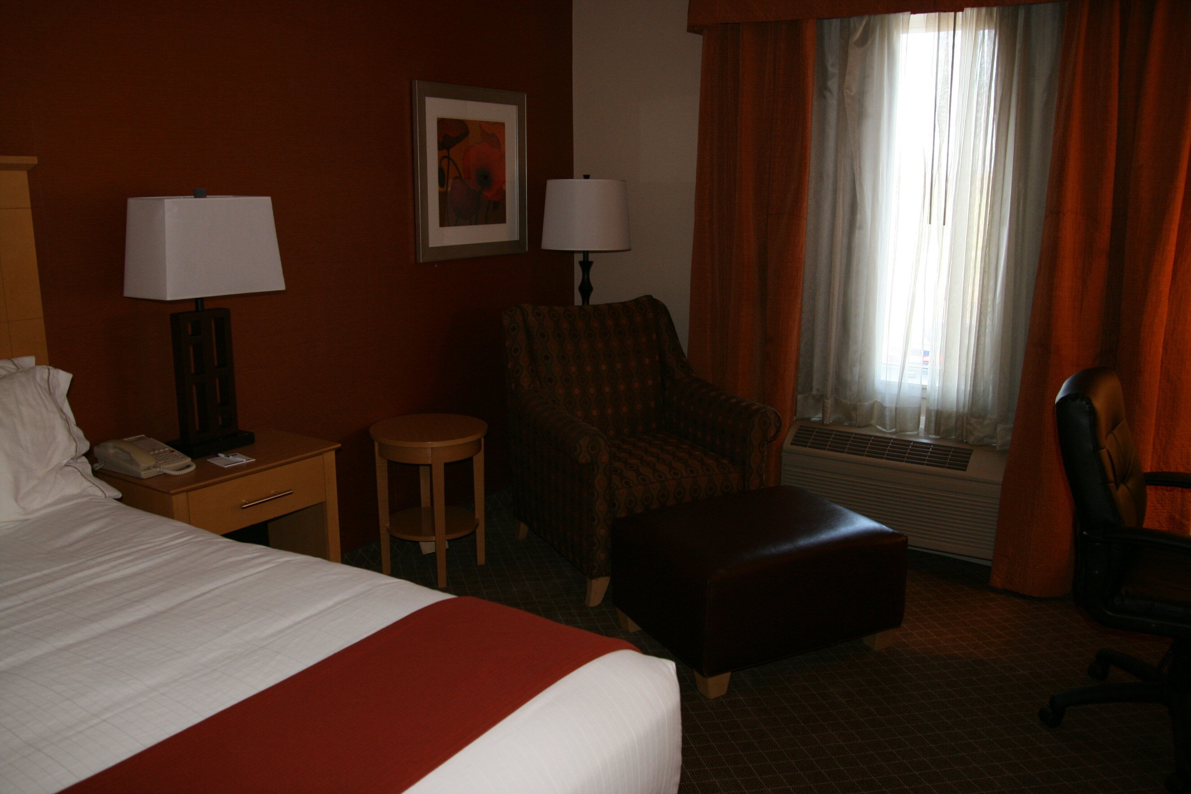 Ein Hotelzimmer mit einem Bett, einem Sessel, einem Schreibtisch mit Telefon, einer Lampe und einem Fenster mit Vorhängen.