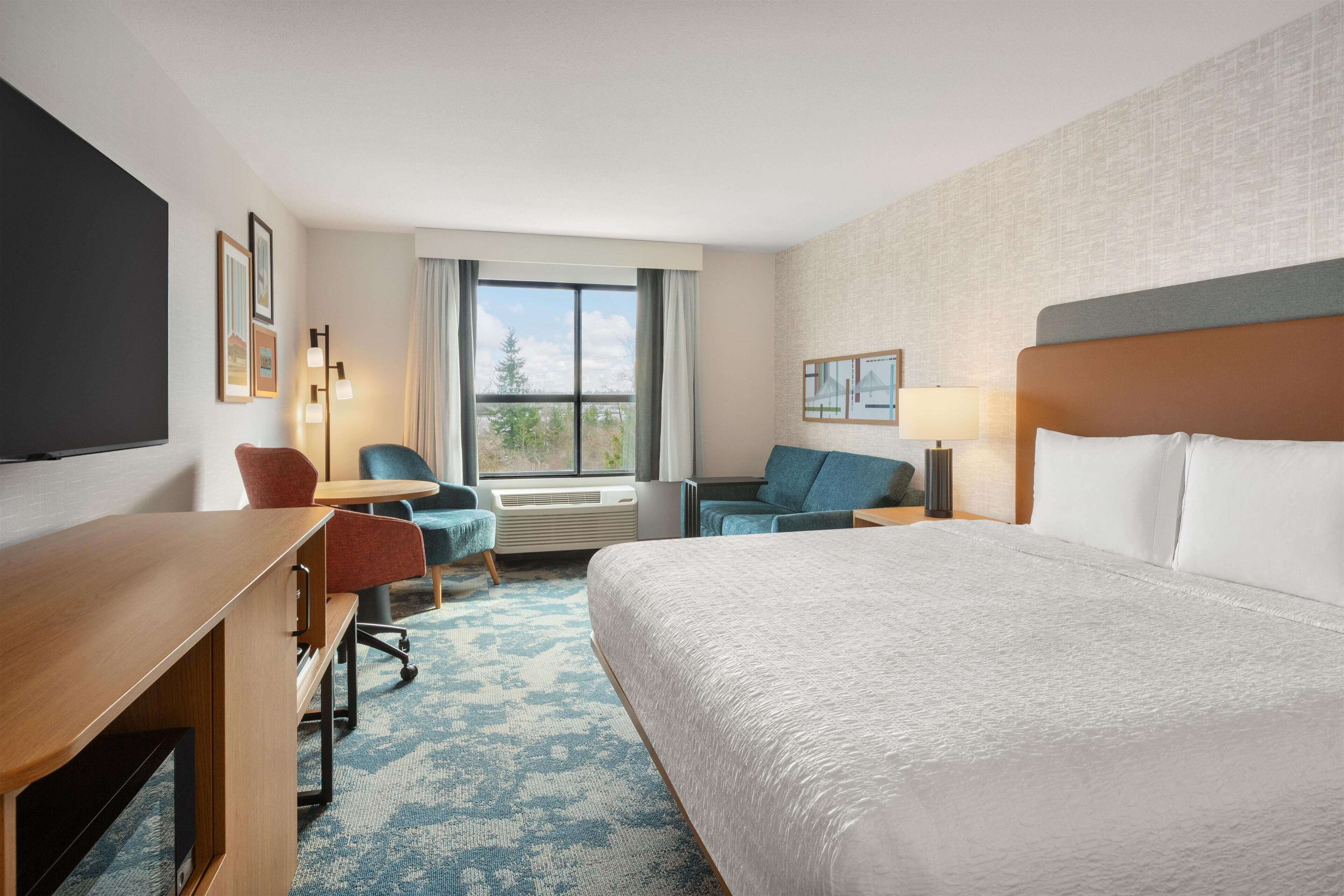 A hotel room with a large bed, a desk, a chair, a small table, and a window with a view of trees.