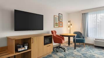 Ein Hotelzimmer mit Fernseher, einem Holfach mit Mikrowelle, einer Kaffeemaschine und einem Tablett mit Tasse und Zubehör. Zwei Stühle, einer rot und einer blau, sowie ein kleiner Tisch sind ebenfalls vorhanden. Der Raum verfügt über einen gemusterten Teppich und ein Fenster mit Vorhängen.