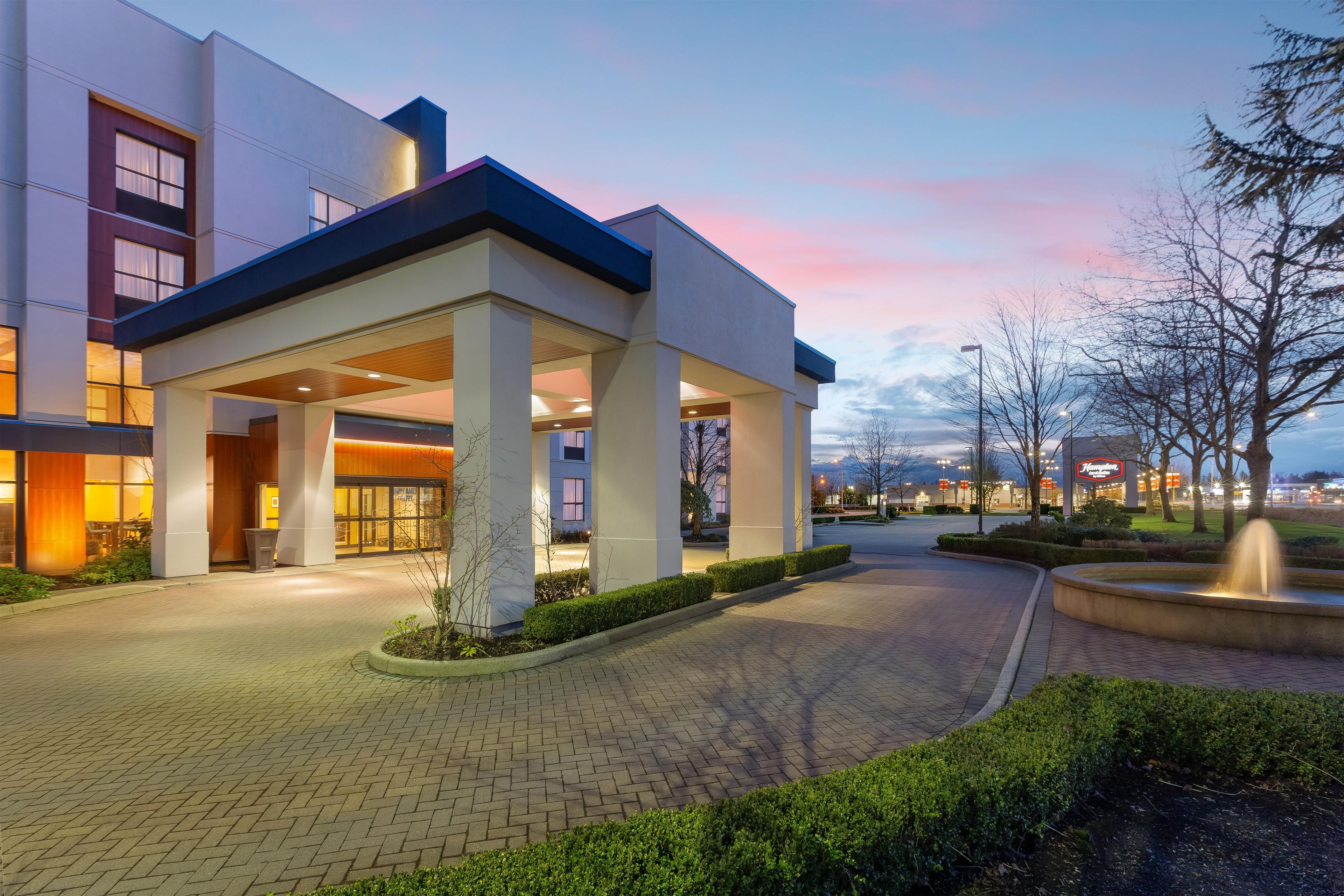 A modern building with a covered entrance, paved walkway, and landscaped area with a fountain.