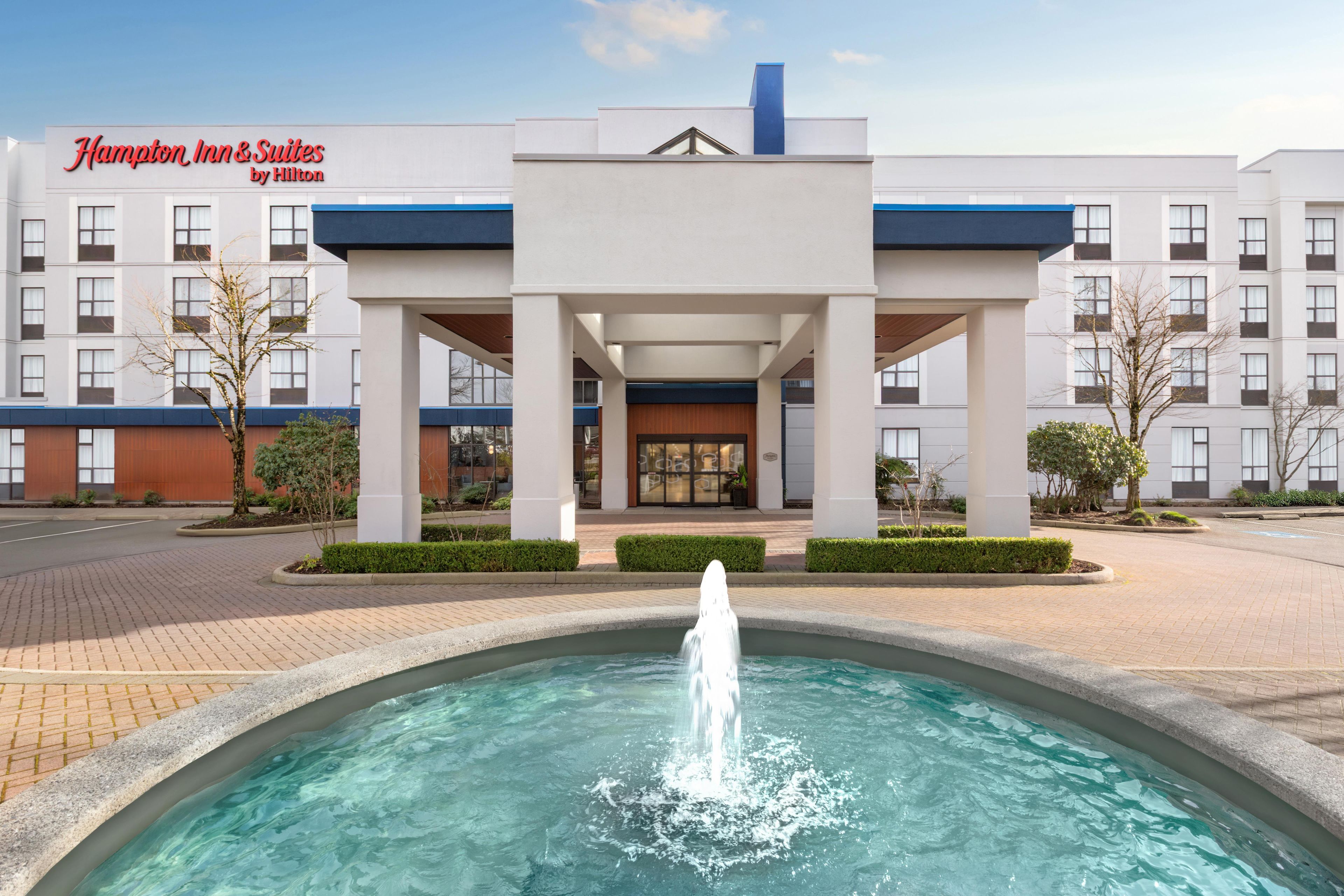 A hotel entrance with a fountain, a clear sky, and a sign reading 'Hampton Inn & Suites by Hilton'.