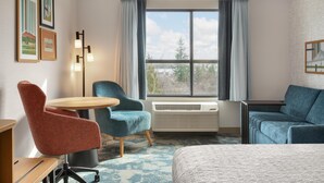 Habitación de hotel con cama, dos sillones, una mesa redonda, un escritorio, una lámpara de pie, una ventana con cortinas y cuadros en la pared.