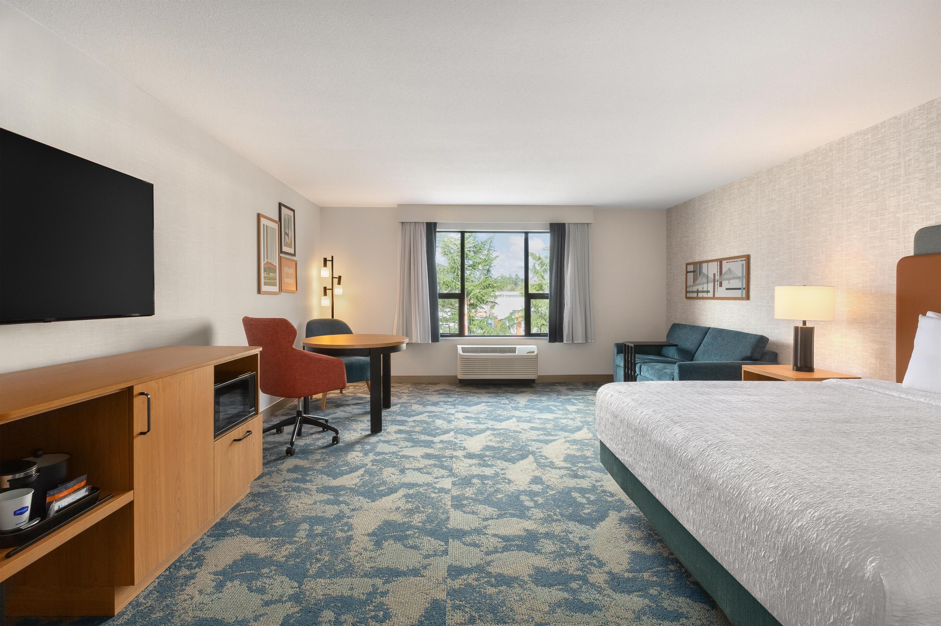A hotel room with a bed, a desk, a chair, a TV, and a window with curtains.