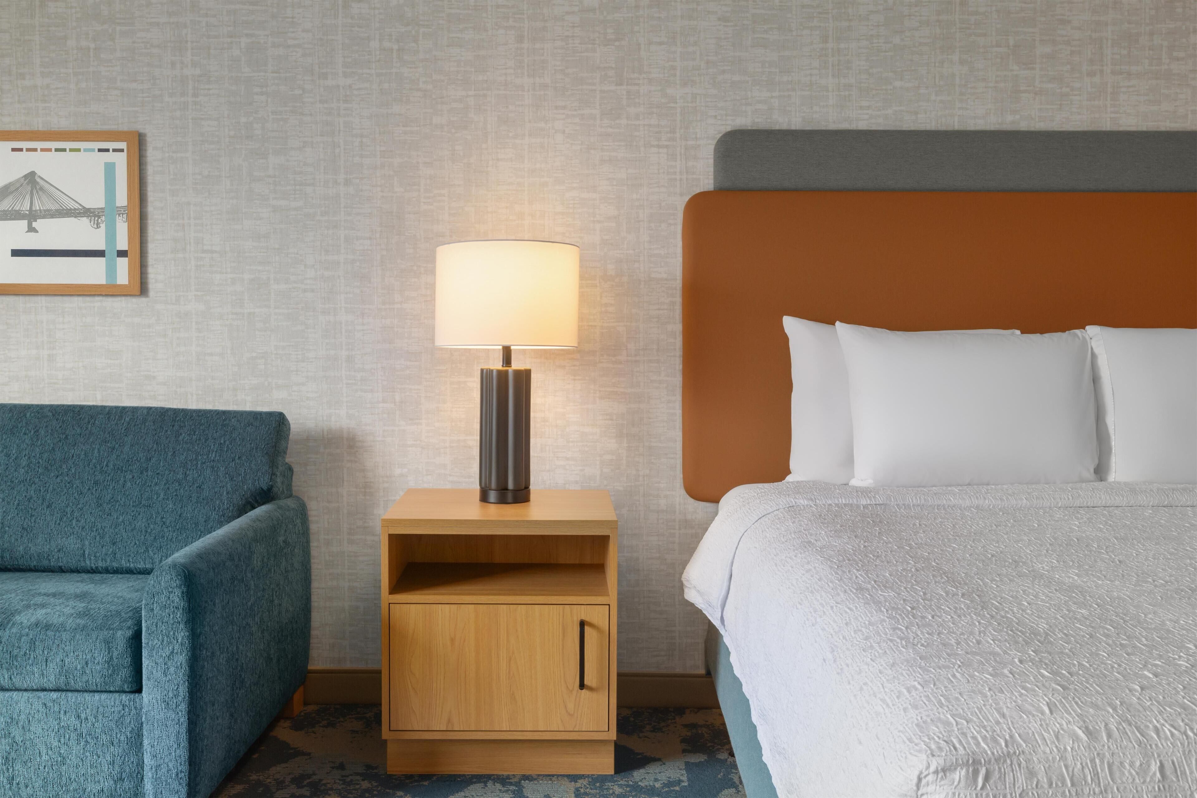 Habitación de hotel con una cama, una mesita de noche con lámpara, un sofá y un cuadro en la pared.