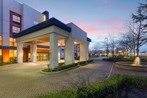 A modern building with a covered entrance, paved pathways, and a landscaped area with a fountain. - Hampton Inn & Suites by Hilton Langley-Surrey (Surrey)