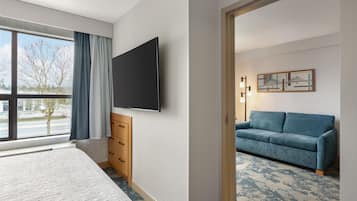 A hotel room with a bed, a flat-screen TV, a dresser, a window with curtains, a sofa, and a patterned carpet.