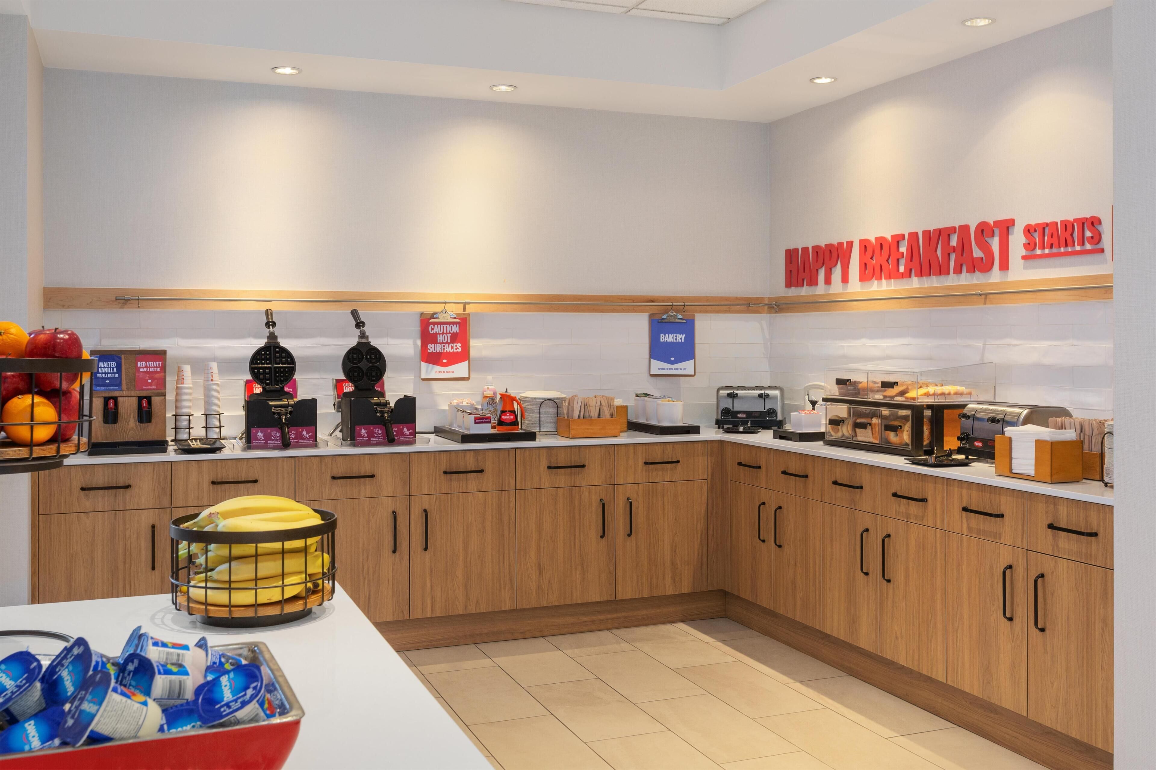 A breakfast area with a sign reading 'HAPPY BREAKFAST STARTS,' a waffle maker, a coffee machine, and a fruit bowl.