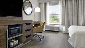 Habitación de hotel con televisión de pantalla plana, escritorio con silla, cama, ventana con cortinas y aire acondicionado.