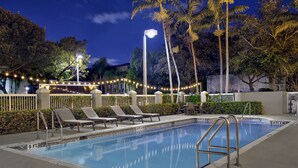 Una piscina con sillas de descanso y una cerca, rodeada de palmeras y luces colgantes.