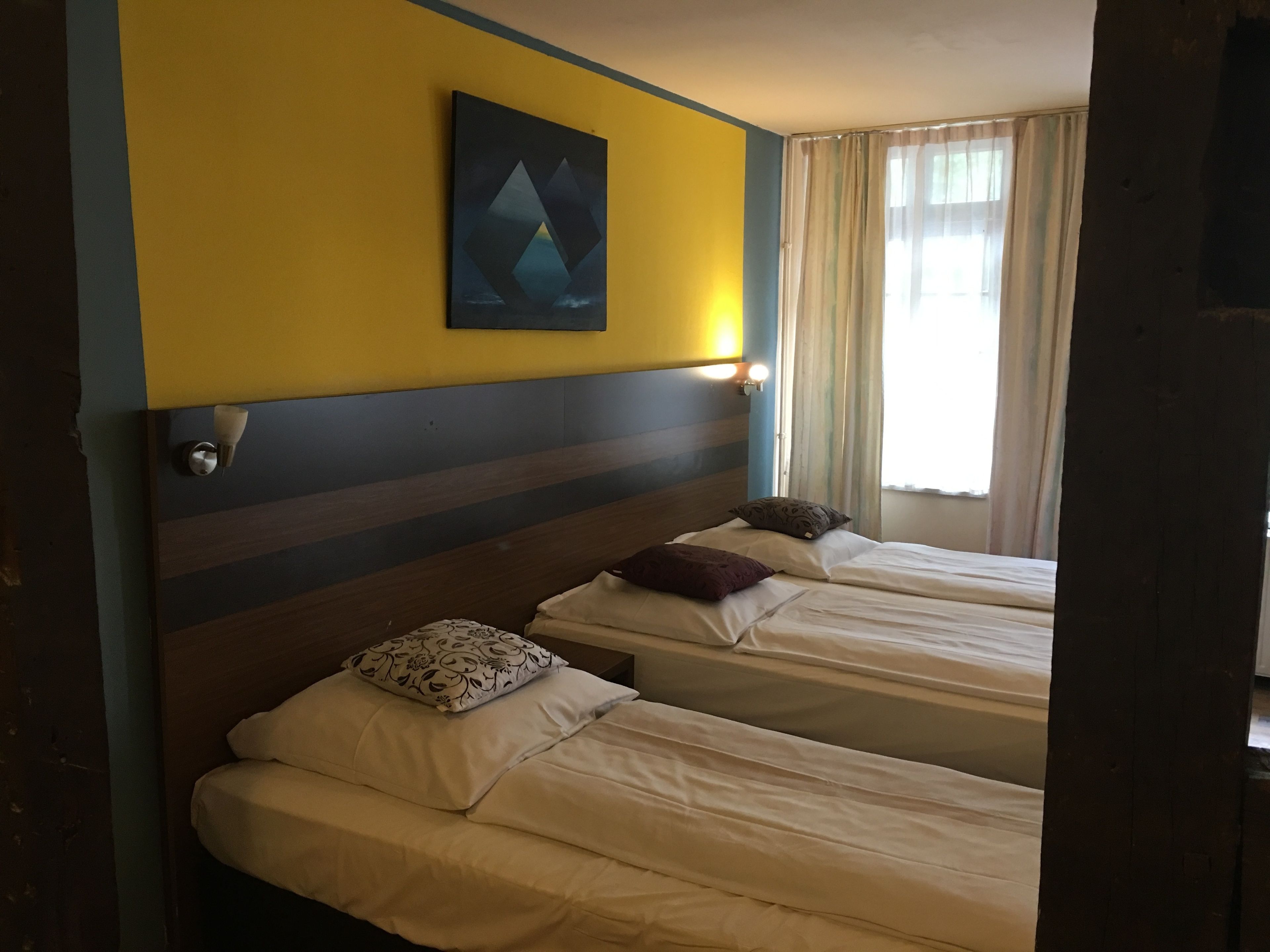 A hotel room with two single beds, a yellow wall, a painting, and a window with curtains.