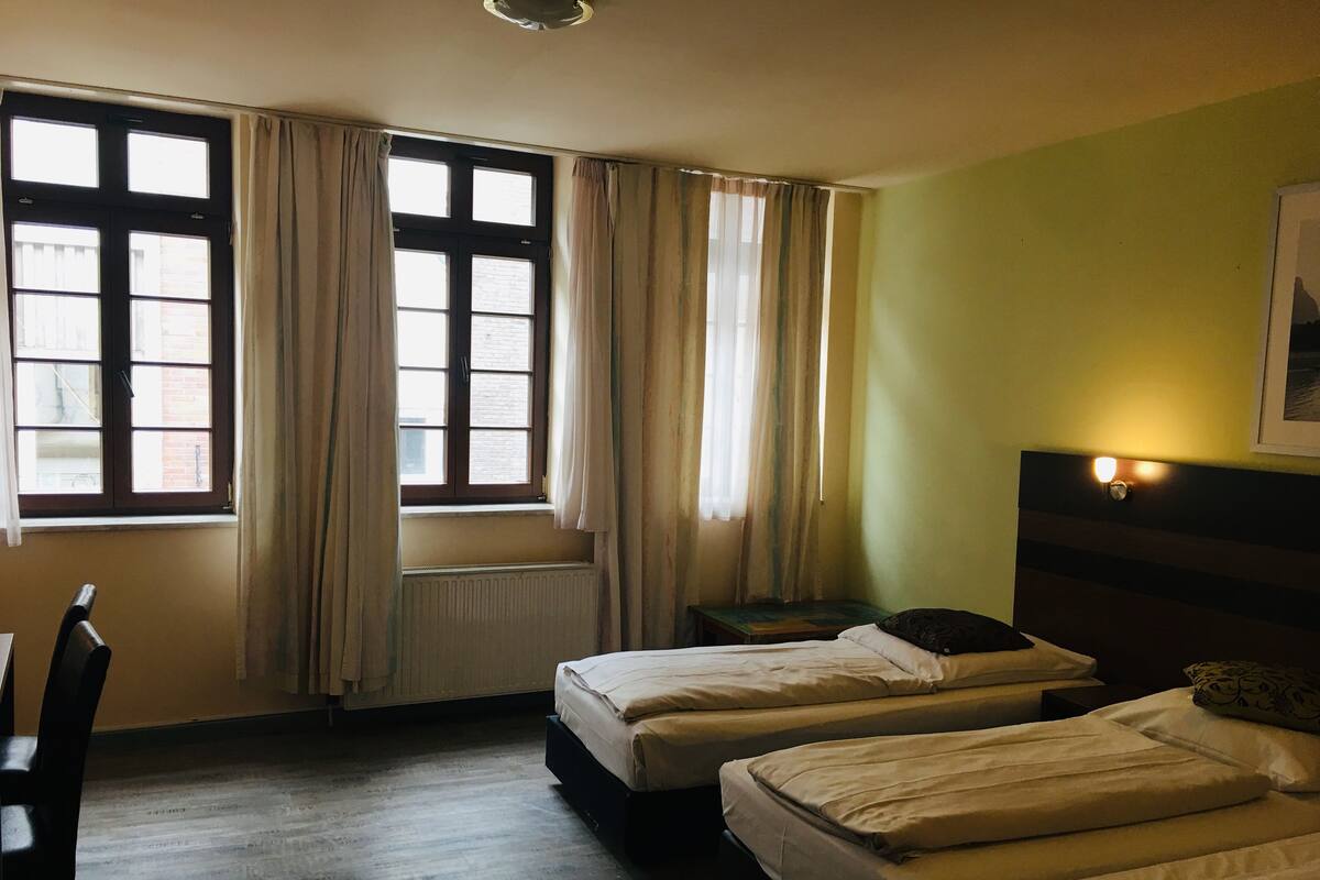 A hotel room with two beds, a chair, a radiator, and a window with curtains.