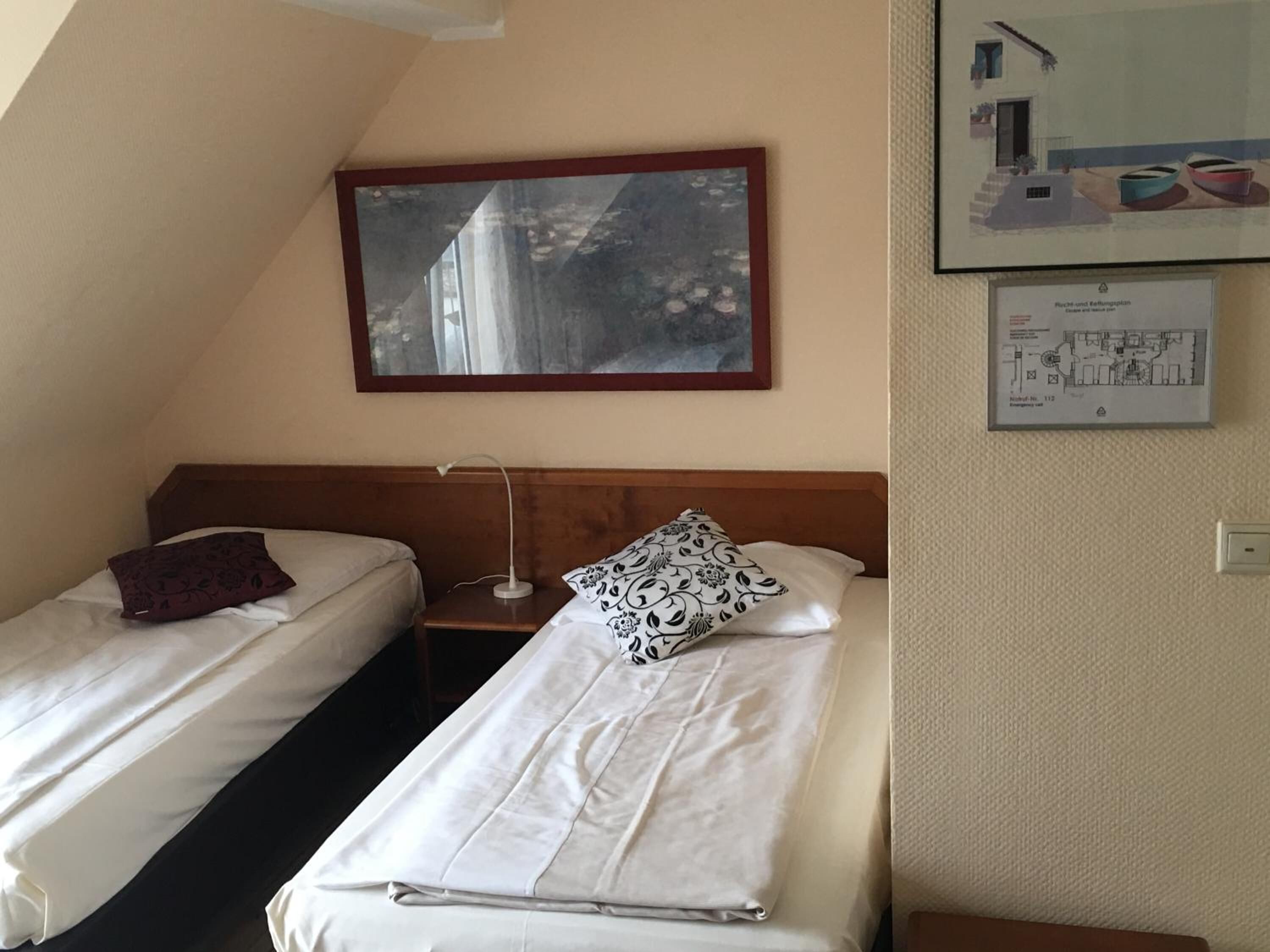 Quarto de hotel com duas camas de solteiro, uma mesa de cabeceira, um abajur, um quadro na parede e um mapa do piso na parede.