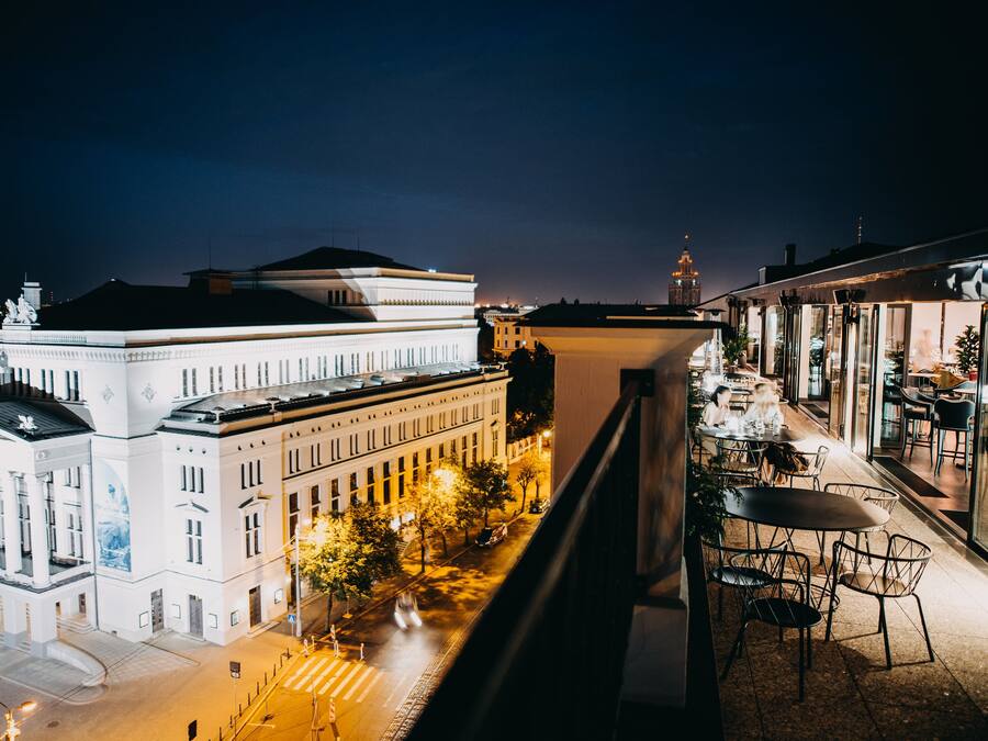 Vista nocturna de un paisaje urbano con un edificio blanco destacado que tiene columnas y una estatua en el techo, una calle con árboles y coches estacionados, y un restaurante con terraza.