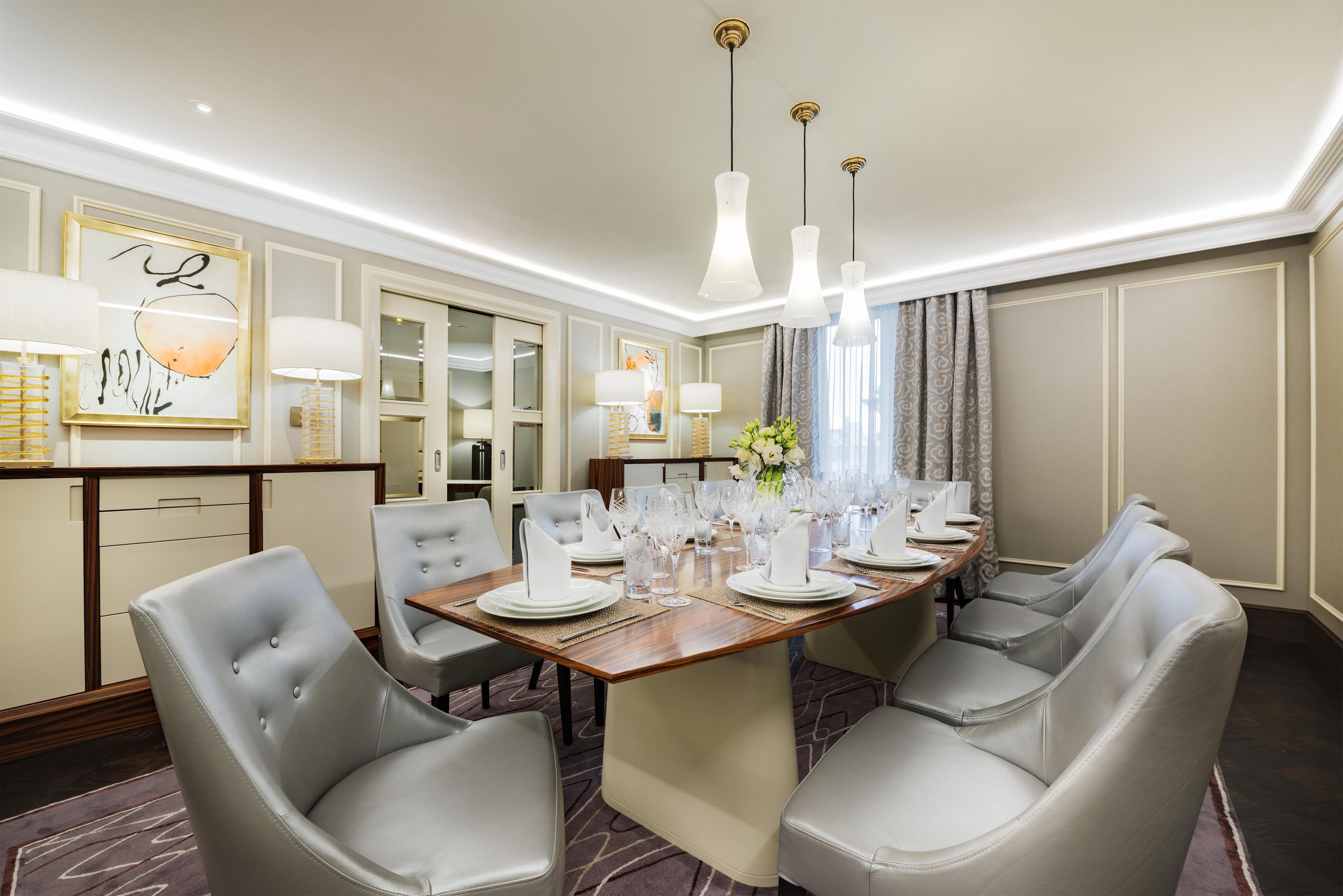 A hotel dining room with a round table set for six, grey upholstered chairs, and pendant lighting.
