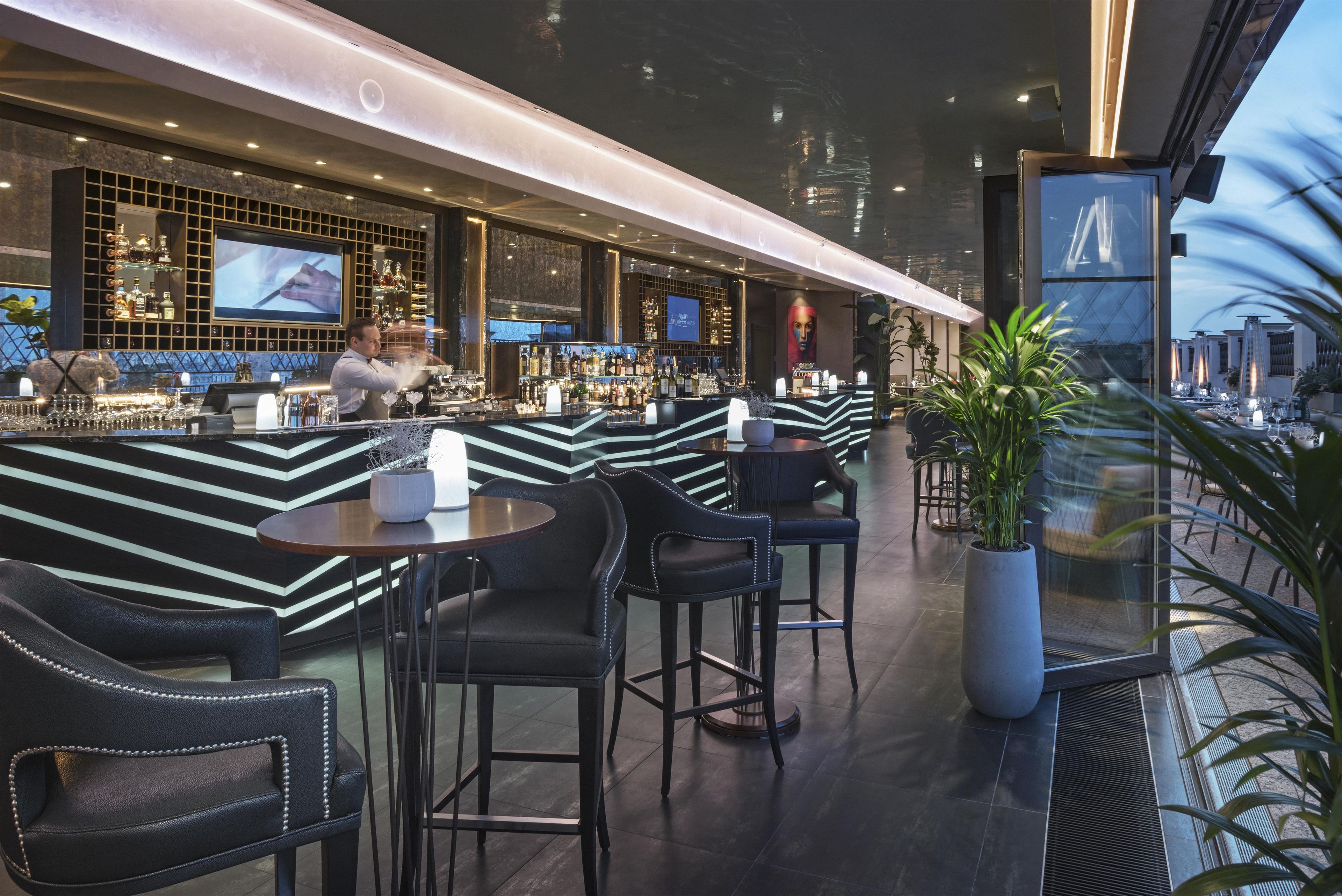 A bar area with a bartender, multiple seating options, and a large screen displaying a program.