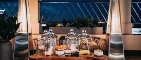 A buffet setup with food items under glass domes, a wooden serving board, and a modern outdoor seating area with metal chairs and potted plants.
