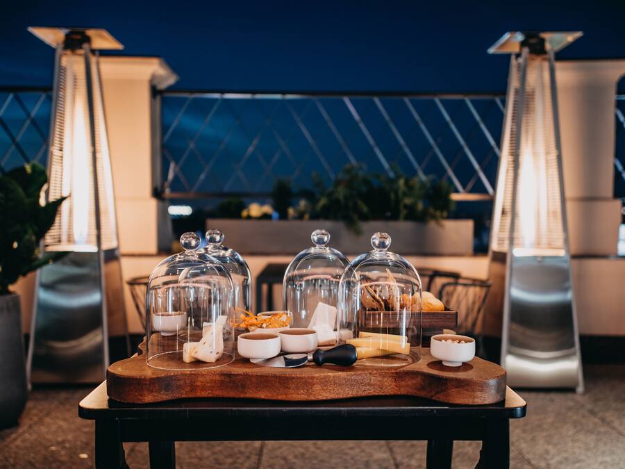 Montaje de bufé con alimentos bajo campanas de cristal, una tabla de madera para servir y un área de estar moderna al aire libre con sillas de metal y plantas en macetas.