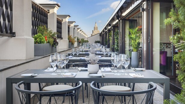 Un área de comedor al aire libre con mesas dispuestas para dos, con un diseño moderno, sillas de metal y una paleta de colores neutros.