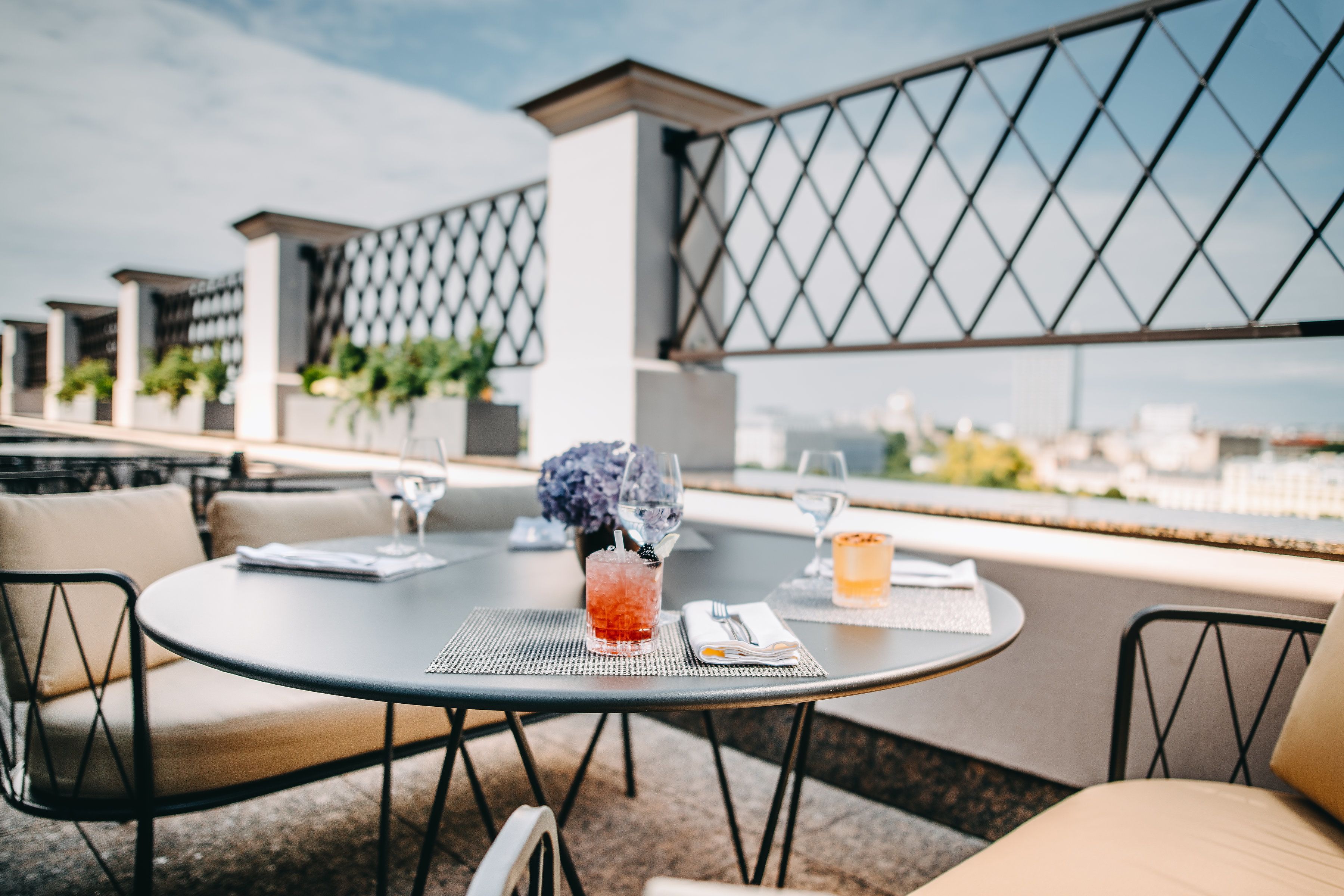 Una mesa redonda con una bebida, un menú y un florero en una terraza con sillas y vistas a edificios.