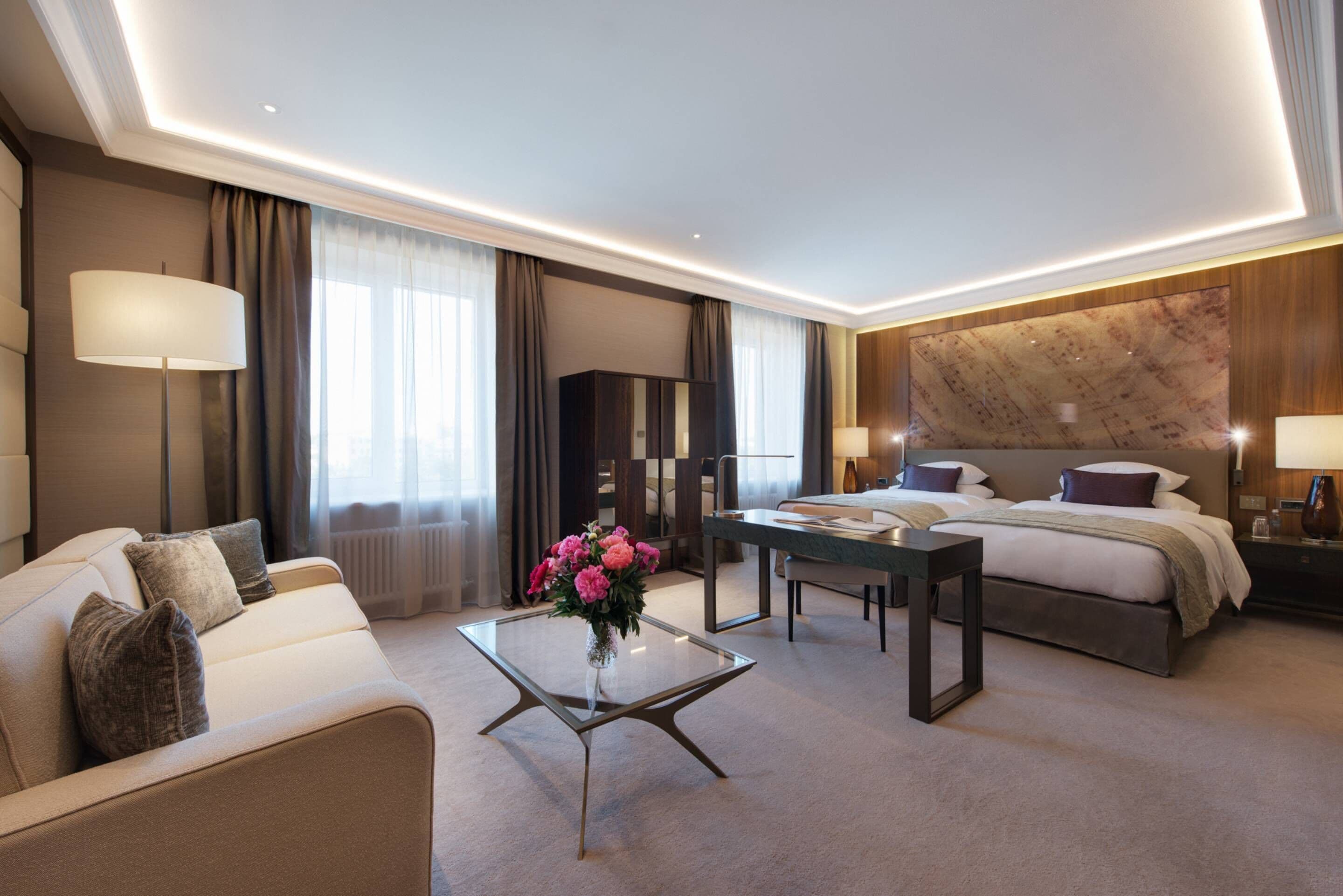 A modern hotel room with a bed, a sofa, a glass coffee table with flowers, a desk, and a large marble wall.