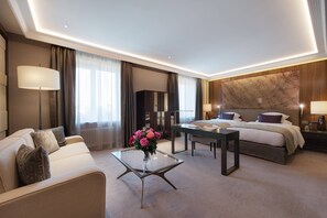 A modern hotel room with a bed, a sofa, a glass coffee table with flowers, a desk, and a large marble wall.