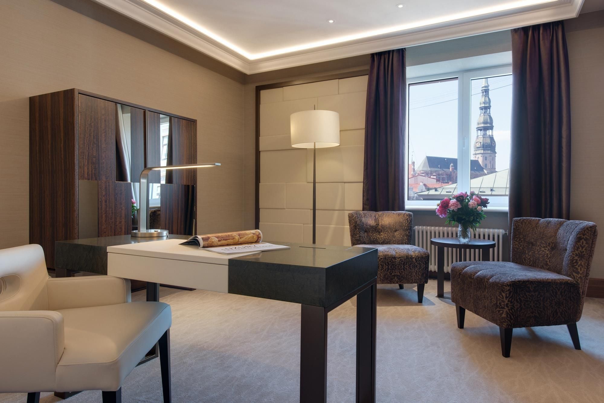 A hotel room with a desk, two chairs, a lamp, and a view of a cityscape.
