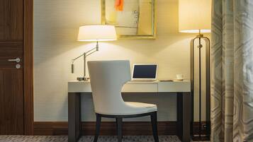 A hotel room with a desk, chair, laptop, two lamps, and a painting on the wall.