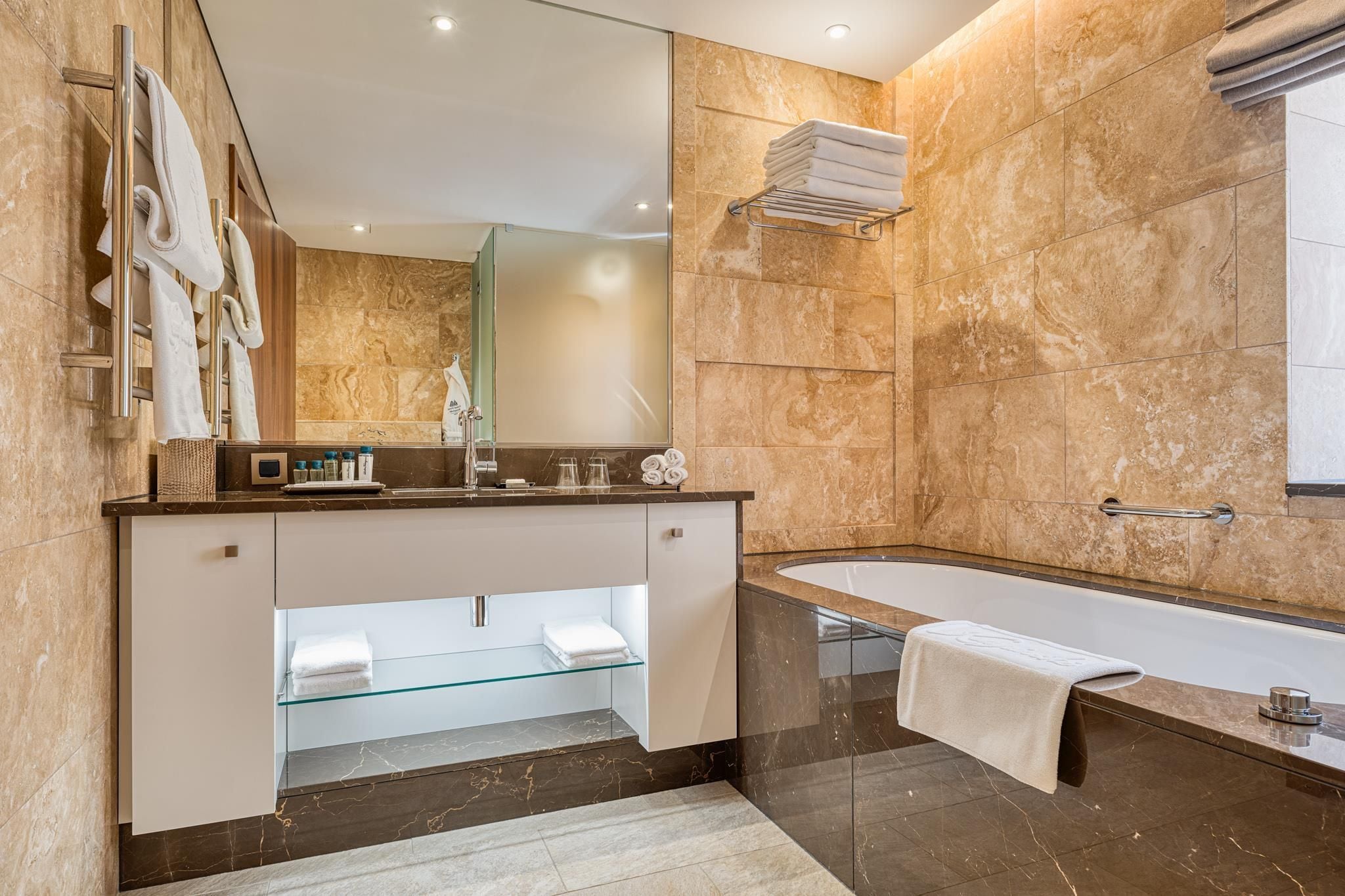 A modern bathroom with a large mirror, a bathtub, and a vanity with a glass shelf.