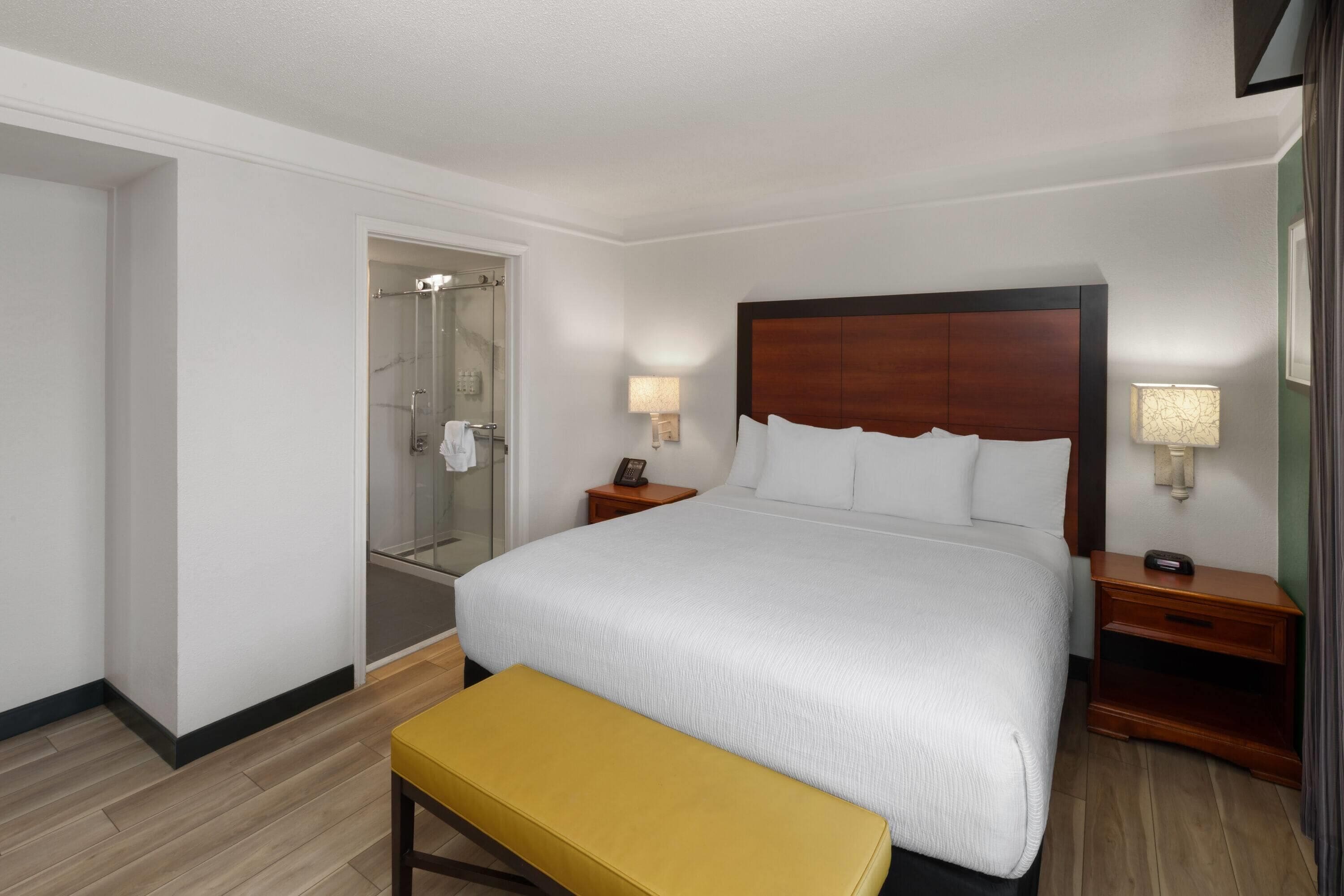 Ein Hotelzimmer mit einem großen Bett, Nachttischen, einer Bank, einer Duschkabine aus Glas und einer Tür zu einem anderen Raum.