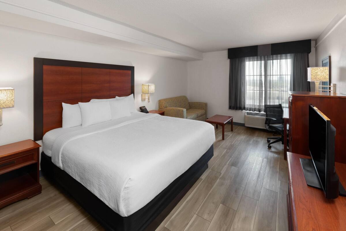 Ein Hotelzimmer mit einem großen Bett, einem Schreibtisch, einem Stuhl, einem Fernseher und einem Fenster mit Vorhängen.