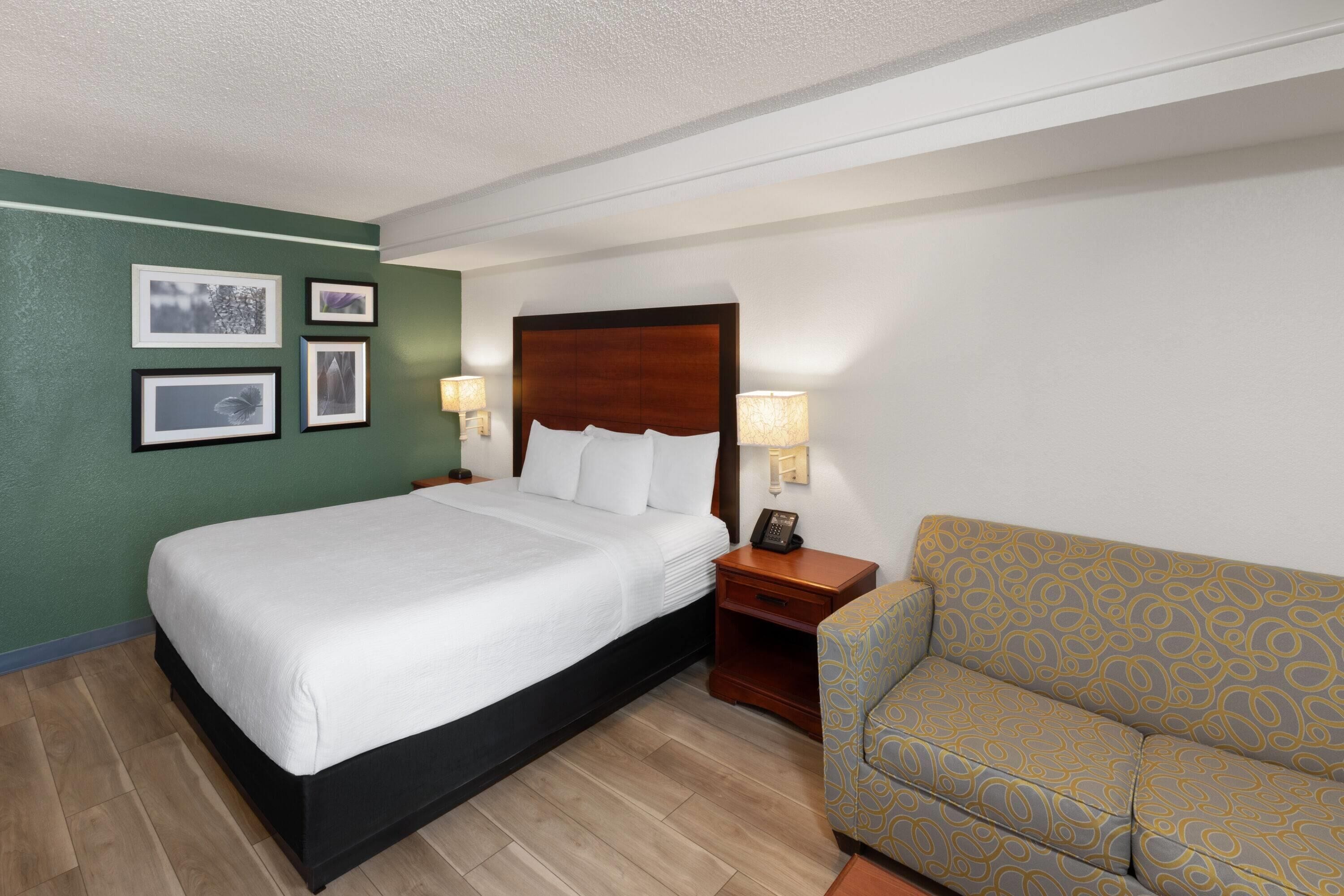 A hotel room with a large bed, a wooden headboard, a nightstand with a lamp, a sofa, and framed pictures on the wall.