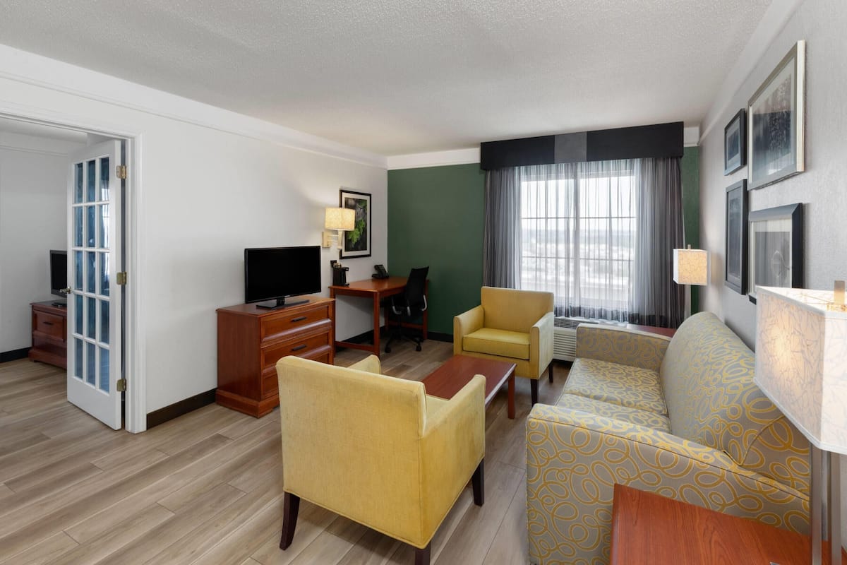 Ein Hotelzimmer mit einer Couch, Sesseln, einem Schreibtisch, einem Fernseher und einem fenster mit Vorhängen.